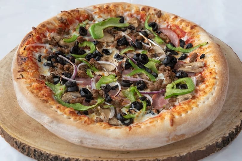 A whole pizza with green peppers, black olives, sausage, mushrooms, and onions on a wooden board.