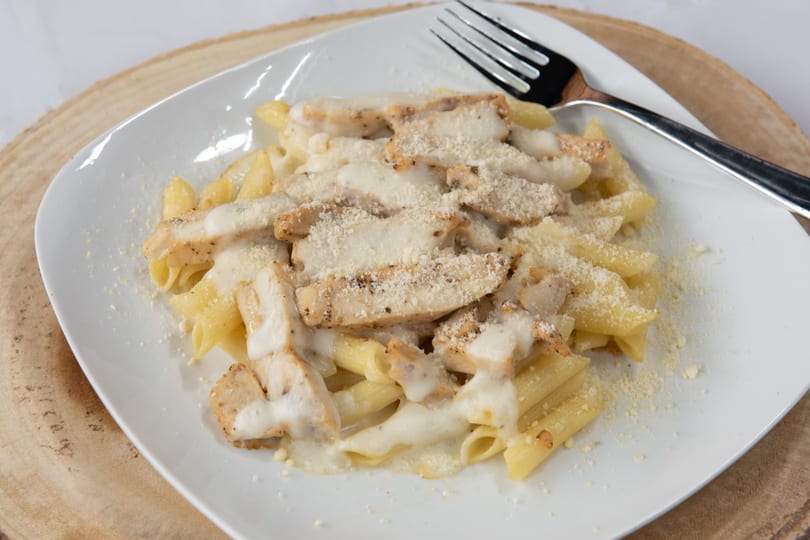 Penne pasta with grilled chicken, creamy white sauce, and grated Parmesan cheese on a white plate.