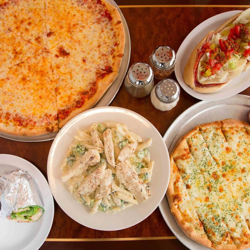 A table laden with a cheese pizza, a sub sandwich, chicken alfredo pasta, garlic knots, and a wrap.