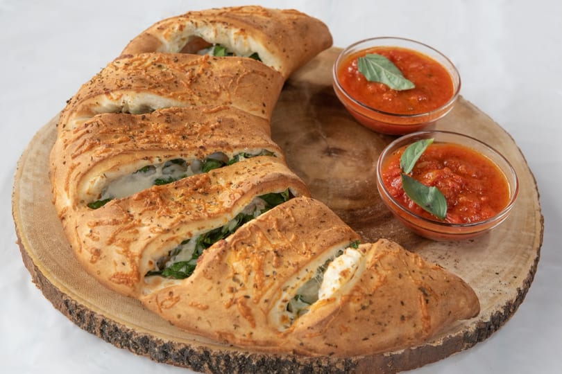 A golden-brown calzone, crescent-shaped and sliced, filled with cheese and spinach, served with two bowls of marinara sauce on a wooden board.